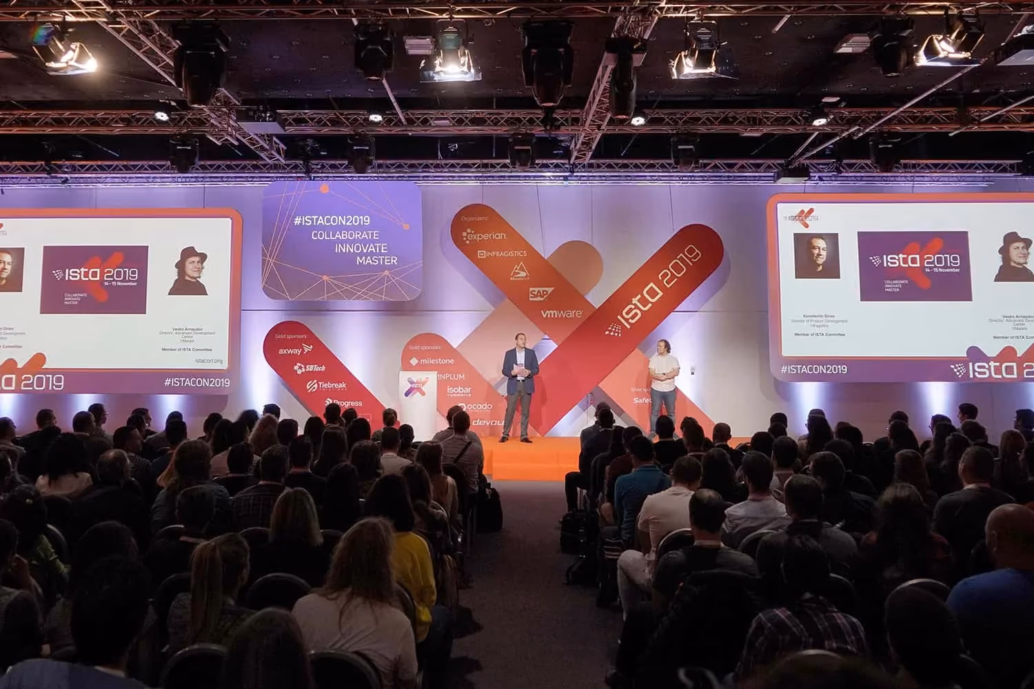 A speaker alone on the ISTA 2019 main stage in front of a packed auditorium, against an orange and purple backdrop reading "#ISTACON2019 Collaborate Innovate Master" flanked by screens of organizer and sponsor logos.