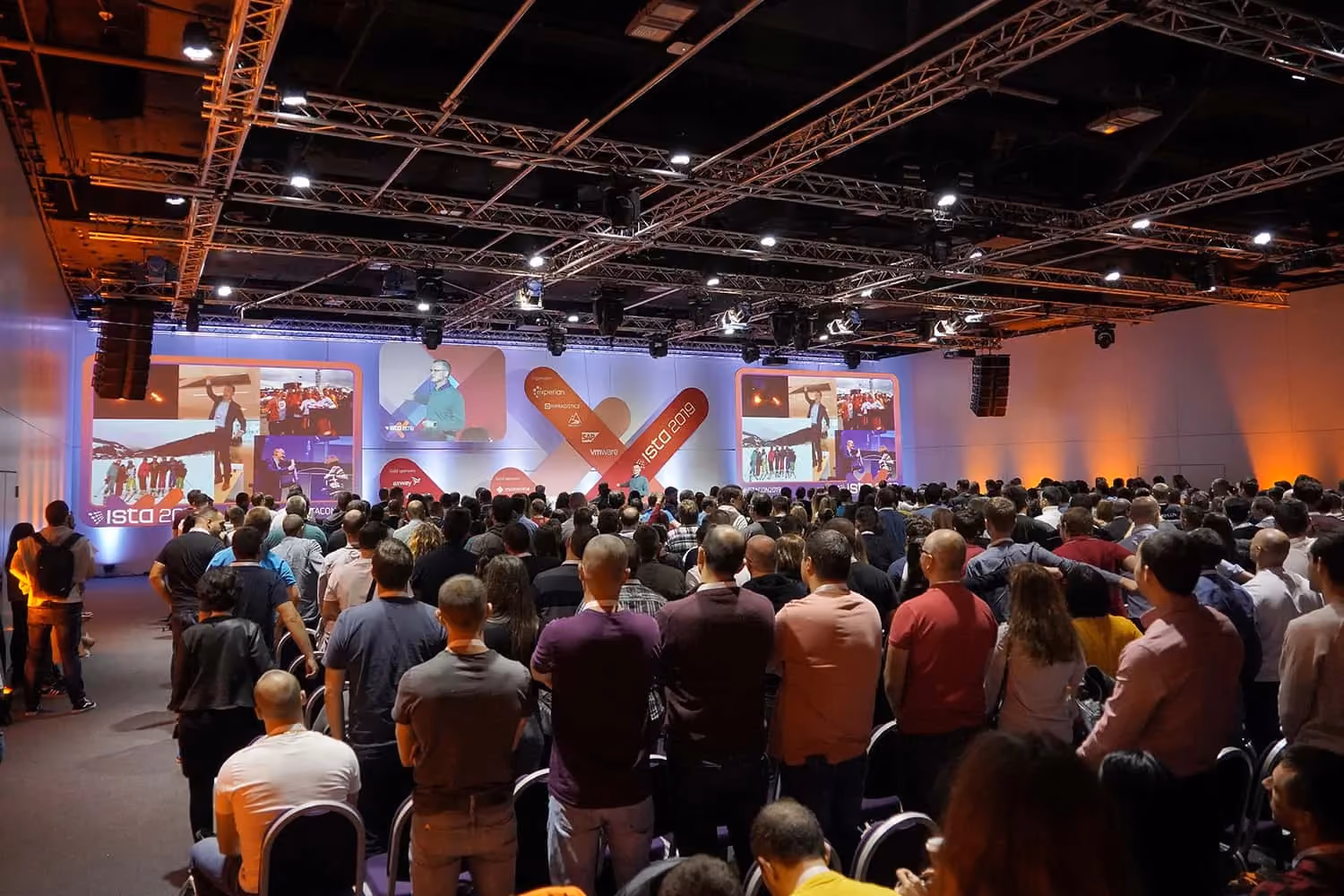 A standing-room-only ISTA 2019 hall seen from the back of the room, hundreds of attendees facing a lit stage with "ISTA 2019" screens and a photo collage on the side displays.