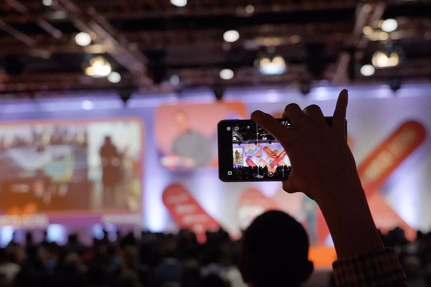 An attendee's raised hand filming the ISTA stage on a phone; the phone screen shows the speaker and the red ISTA banners while the darkened hall behind is out of focus.