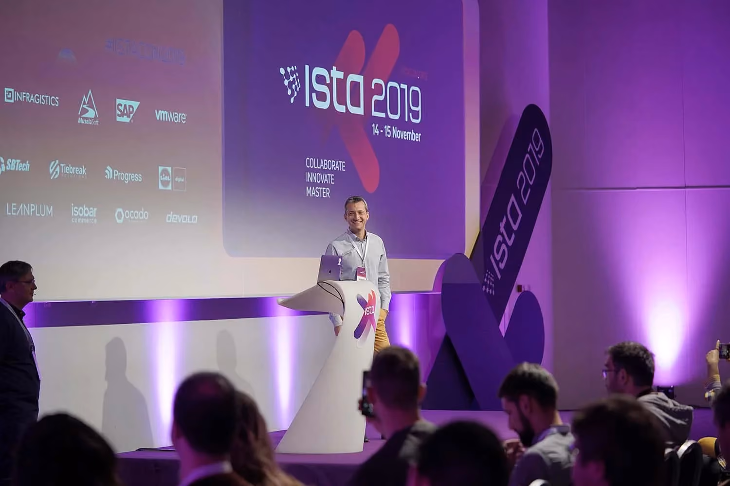 A smiling speaker at a white ISTA lectern on the ISTA 2019 stage, beside a purple screen reading "ISTA 2019, 14 - 15 November, Collaborate Innovate Master" and a grid of sponsor logos.