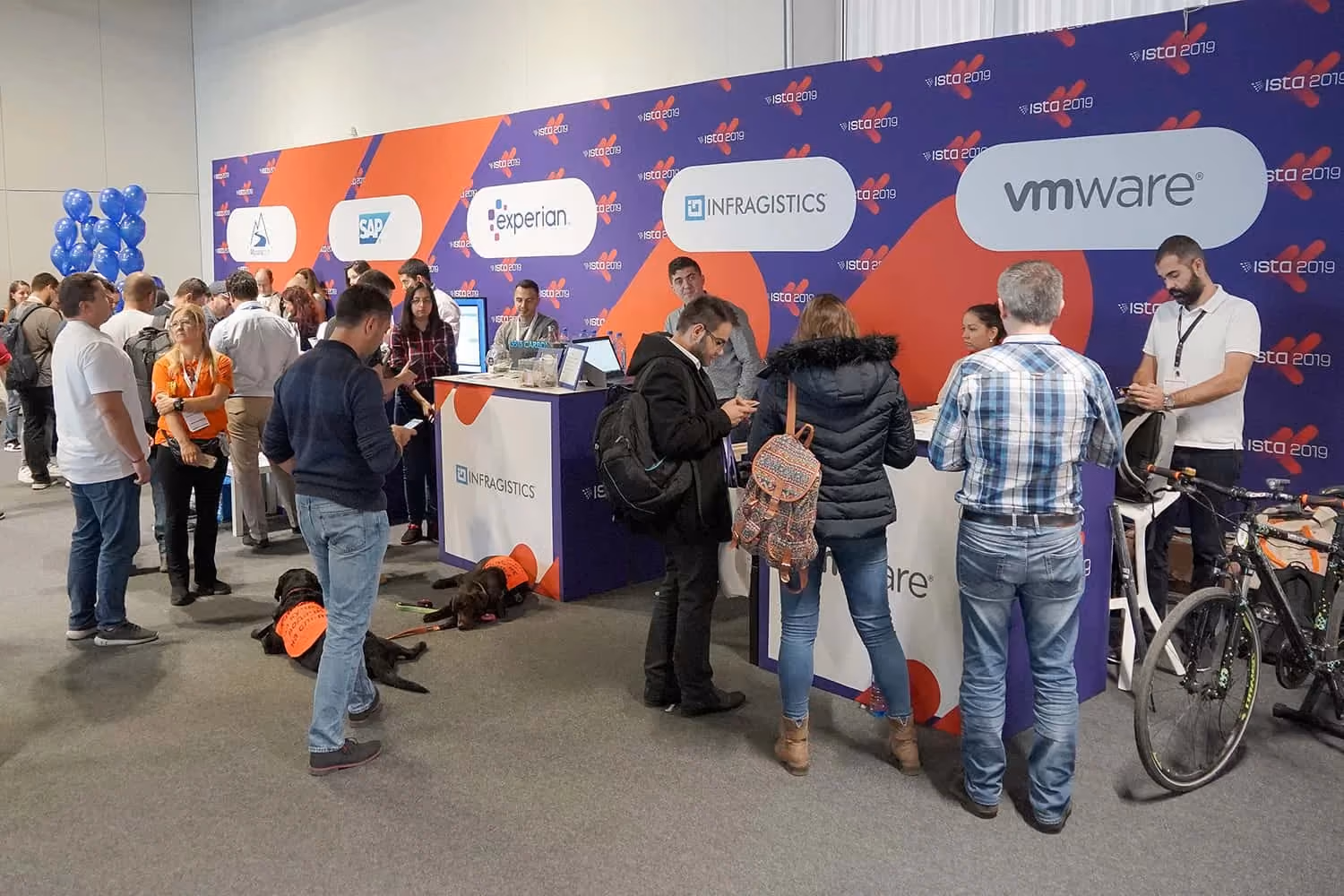 Attendees talking and queueing at the ISTA 2019 registration and expo desks in front of a purple sponsor wall carrying the Experian, Infragistics and VMware logos; two assistance dogs in orange vests lie on the floor and a bicycle stands to the right.