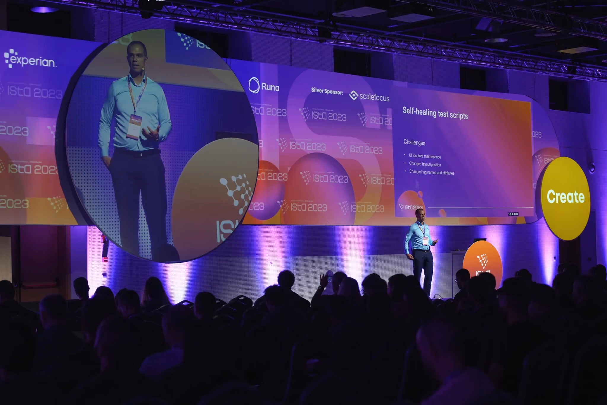 An ISTA 2023 speaker in a light blue shirt presenting a slide titled "Self-healing test scripts", his image relayed on a large circular screen above the stage and a yellow "Create" disc at the right.