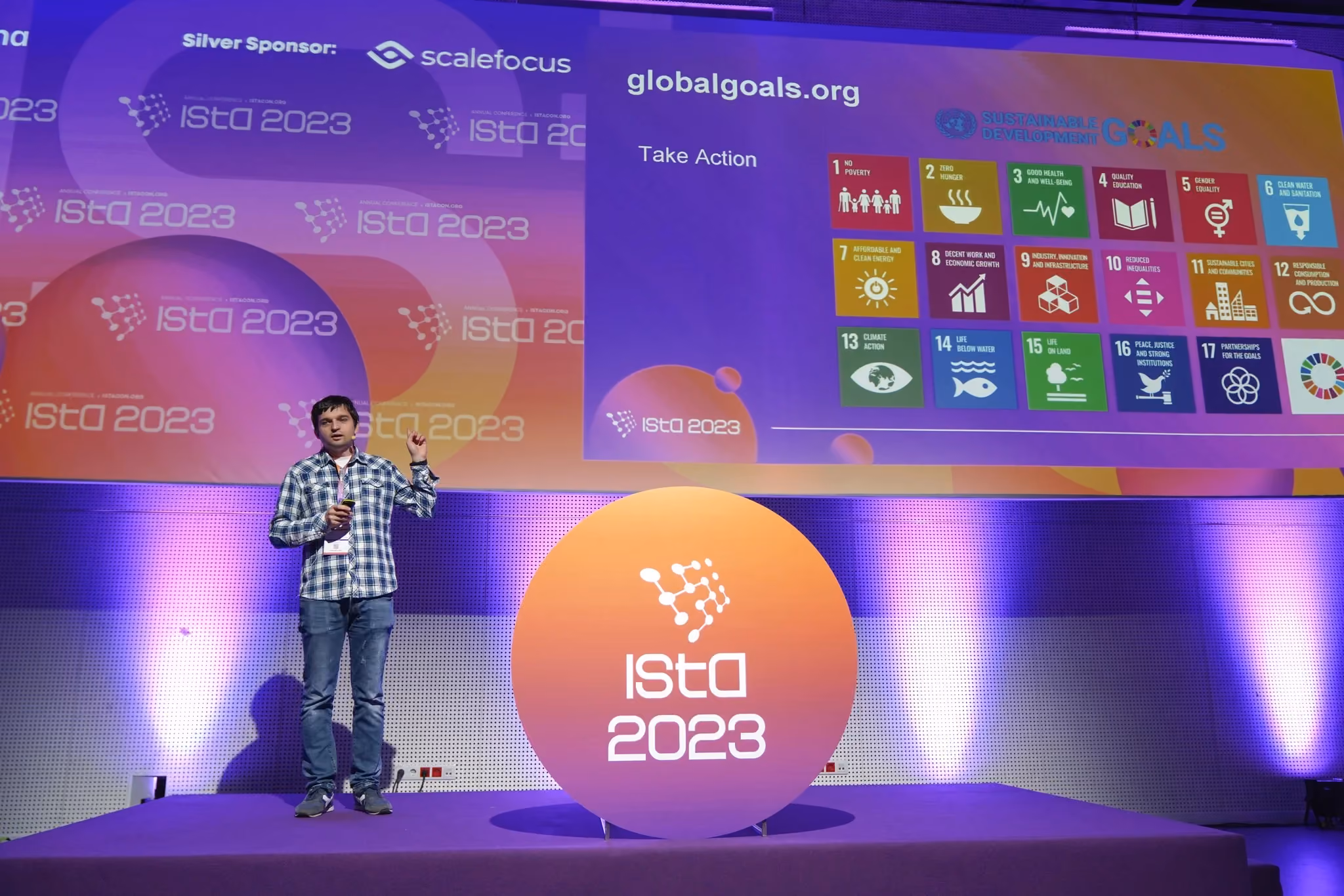 An ISTA 2023 speaker in a checked shirt presenting a slide about globalgoals.org and the UN Sustainable Development Goals, standing beside a large orange "ISTA 2023" disc.