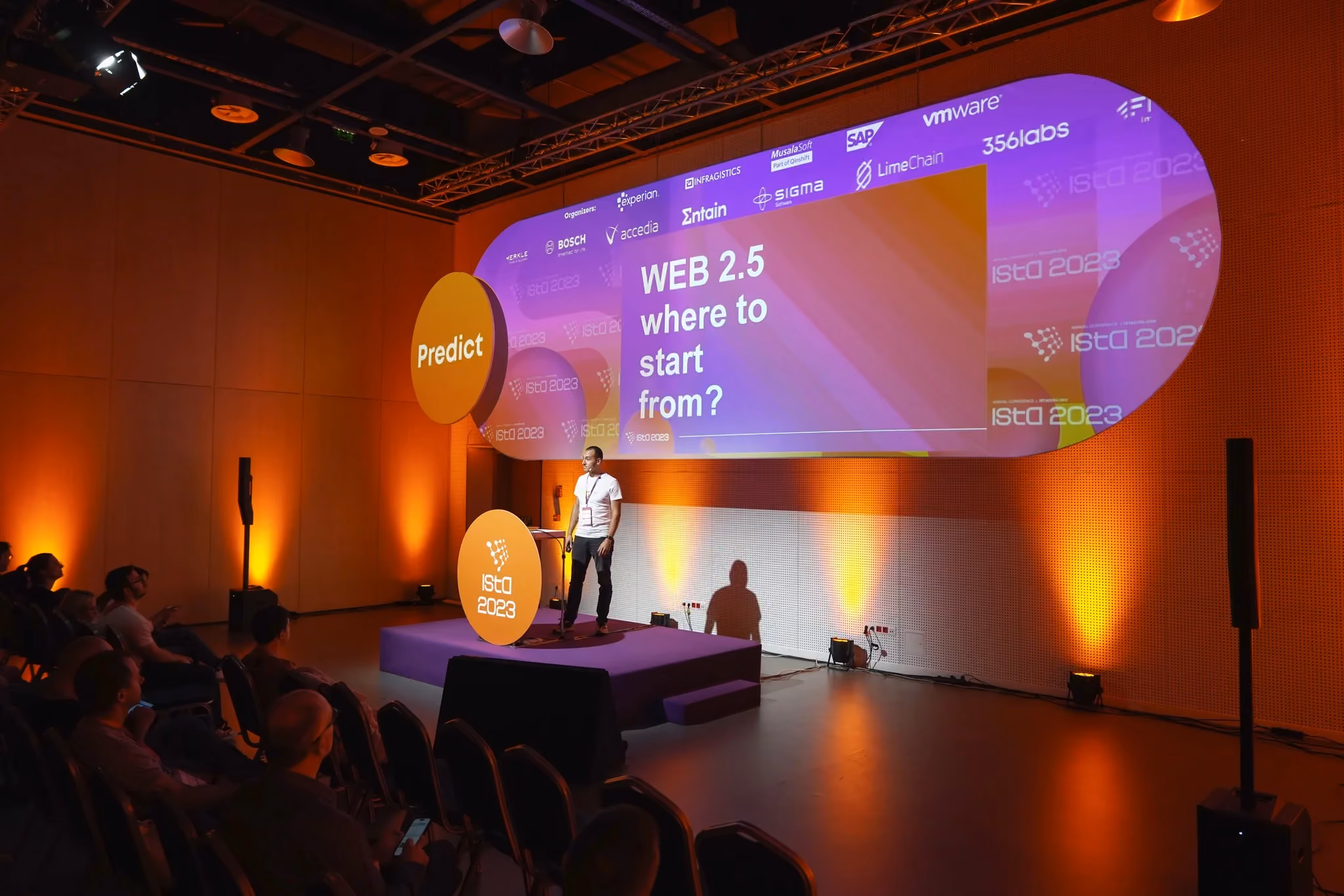 A speaker in a white t-shirt on the ISTA 2023 stage in front of a slide reading "WEB 2.5 where to start from?", with an orange "Predict" disc to the left and seated attendees in the foreground.