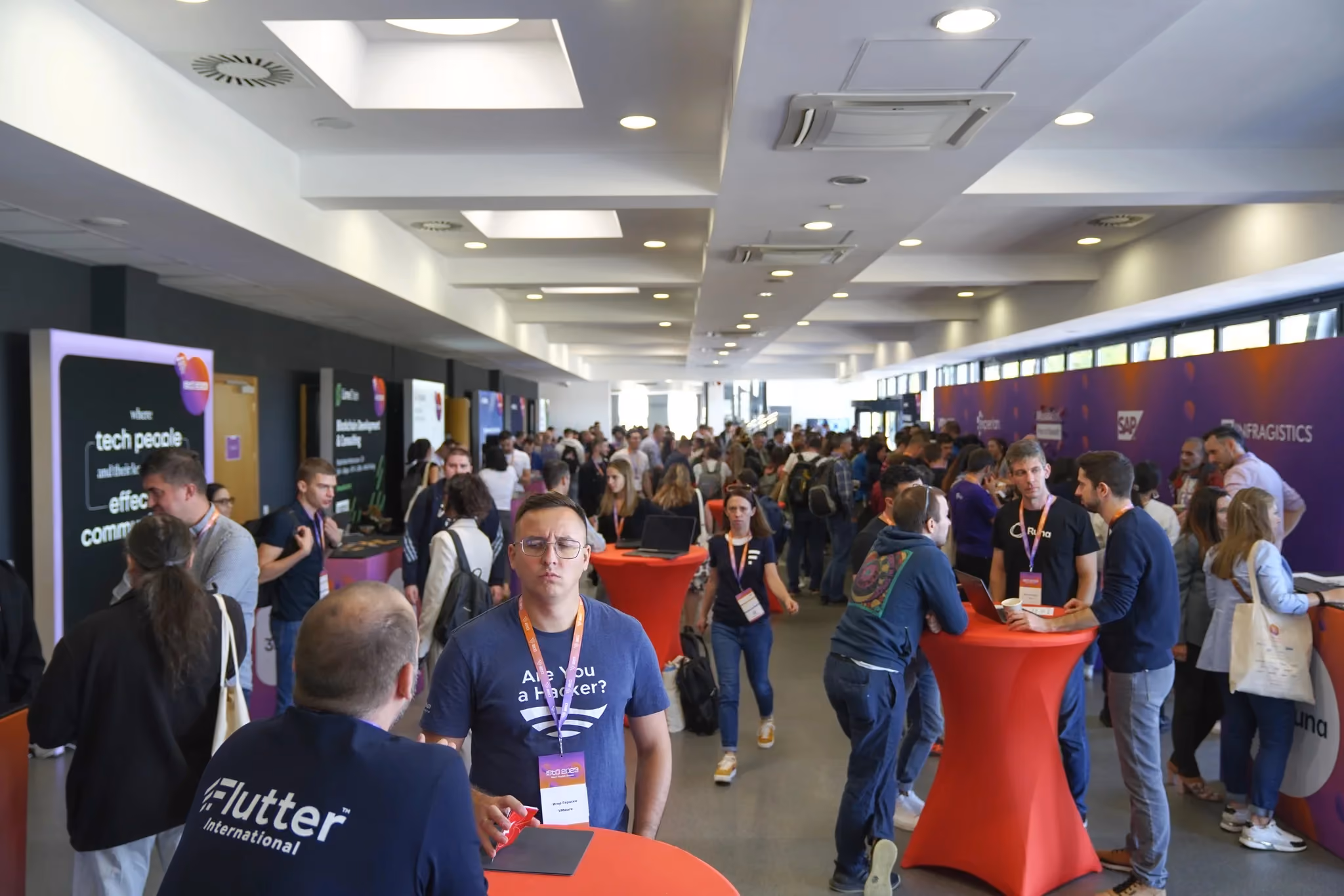 A busy ISTA 2023 networking foyer: attendees in lanyards talking around red cocktail tables between partner stands, with an Infragistics-branded wall along the right.