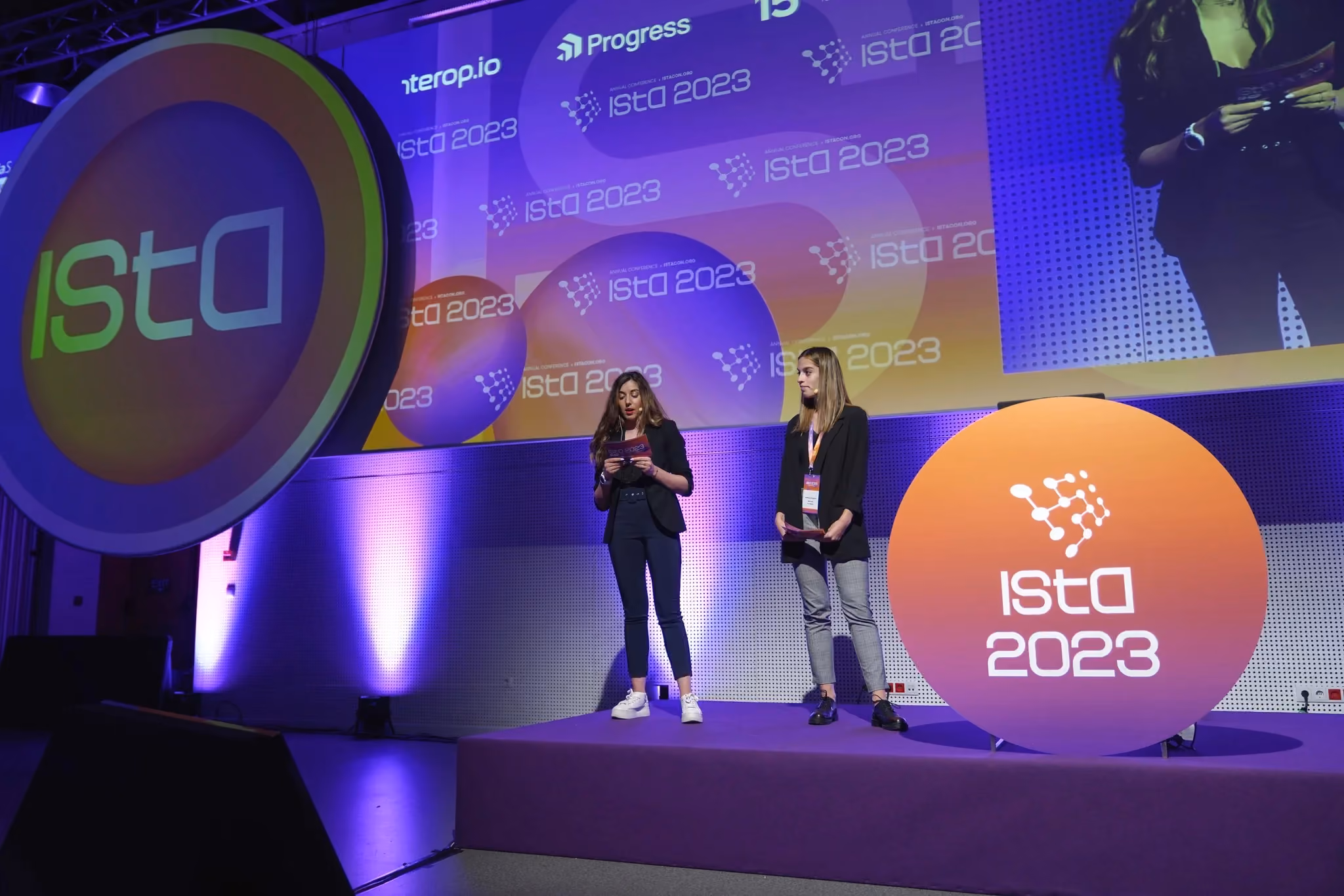 Two hosts in black reading from cards on the ISTA 2023 stage, between a large circular ISTA disc on the left and an orange "ISTA 2023" disc on the right.
