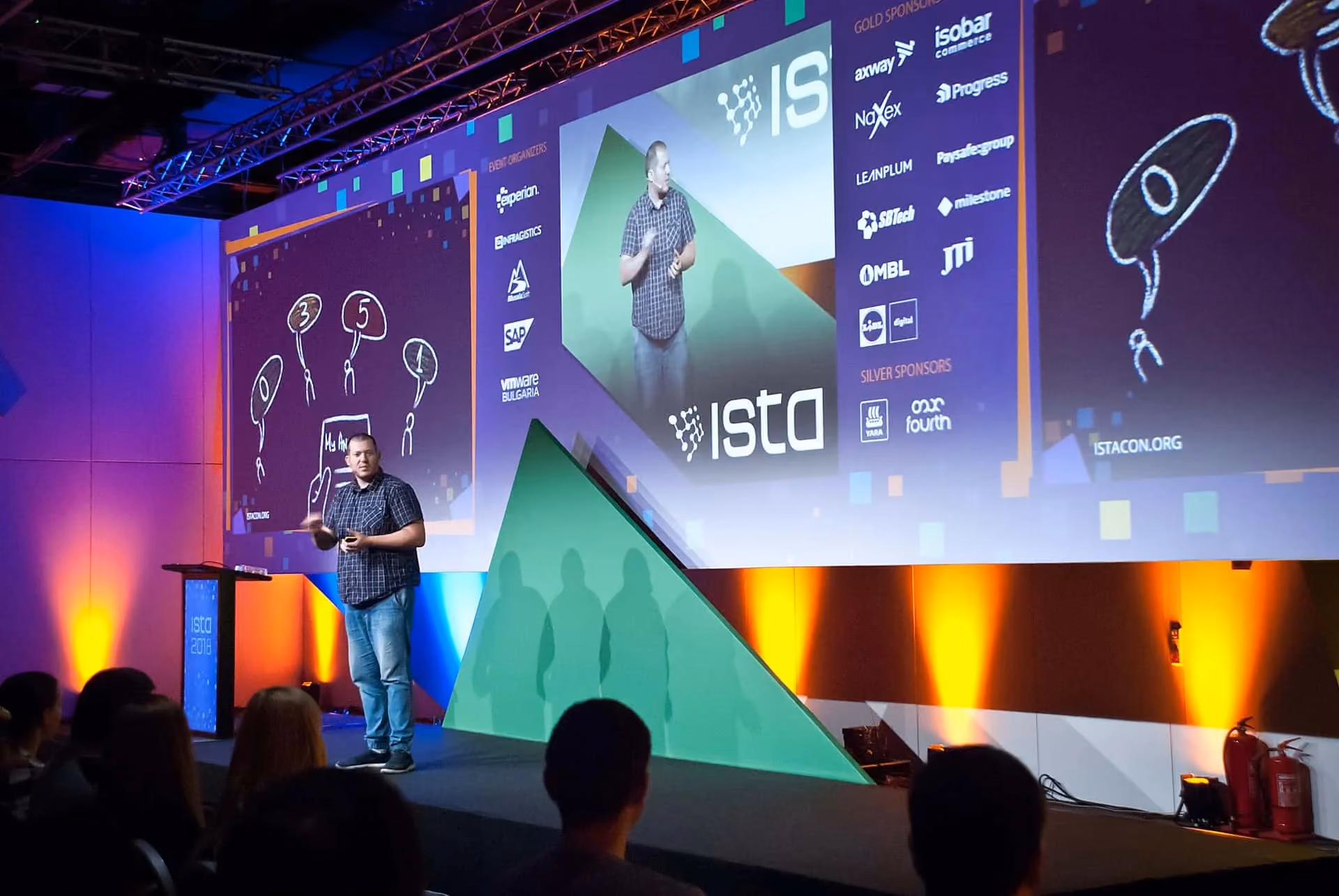 A speaker in a checked shirt on the ISTA 2018 stage in front of a wide screen showing his hand-drawn slide, his live camera feed and columns of event organizer, gold and silver sponsor logos.
