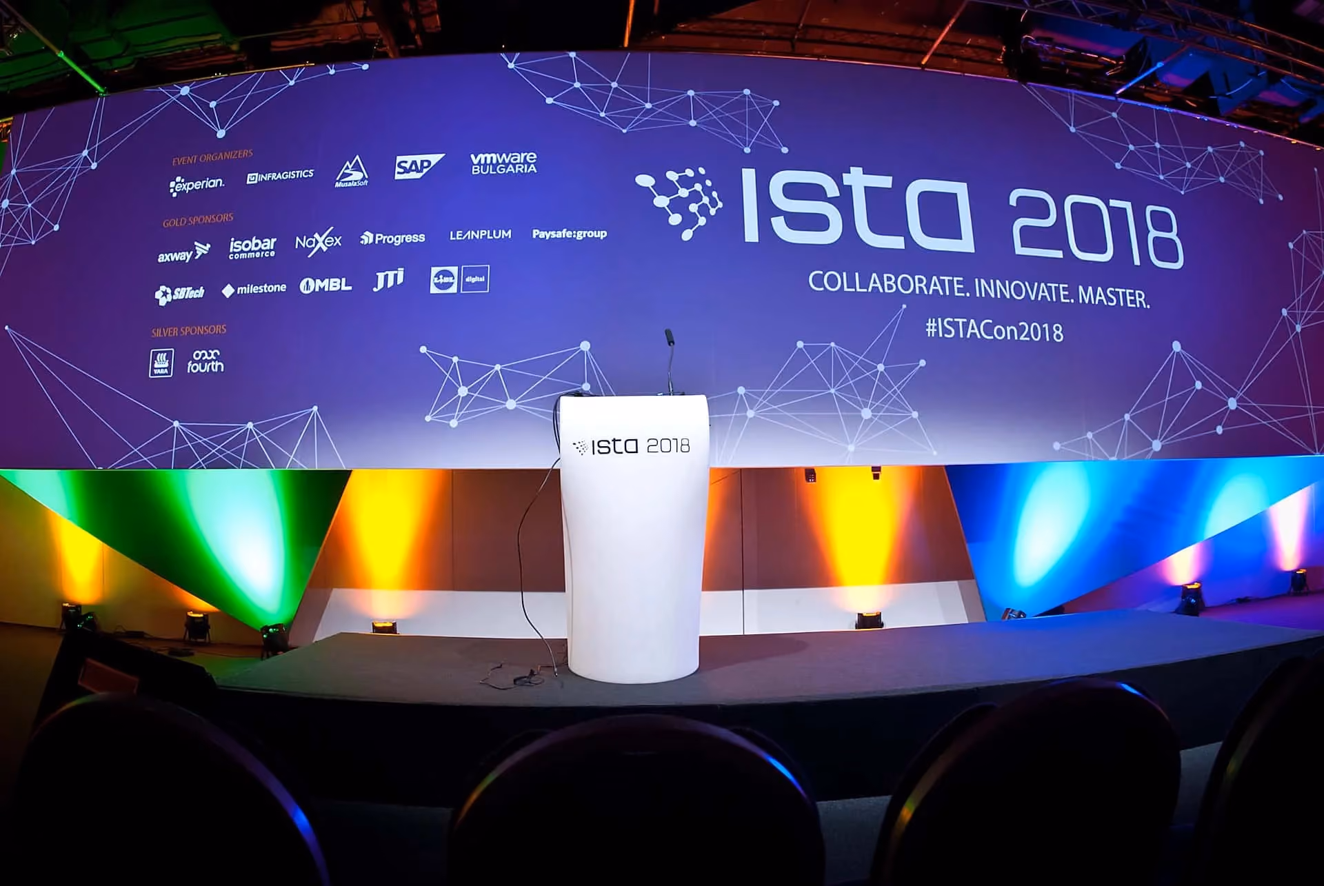 The empty ISTA 2018 stage before the sessions start: a white ISTA lectern and microphone in front of a purple backdrop reading "ISTA 2018 — Collaborate. Innovate. Master. #ISTACon2018" above rows of organizer and sponsor logos, lit by coloured floor lights.