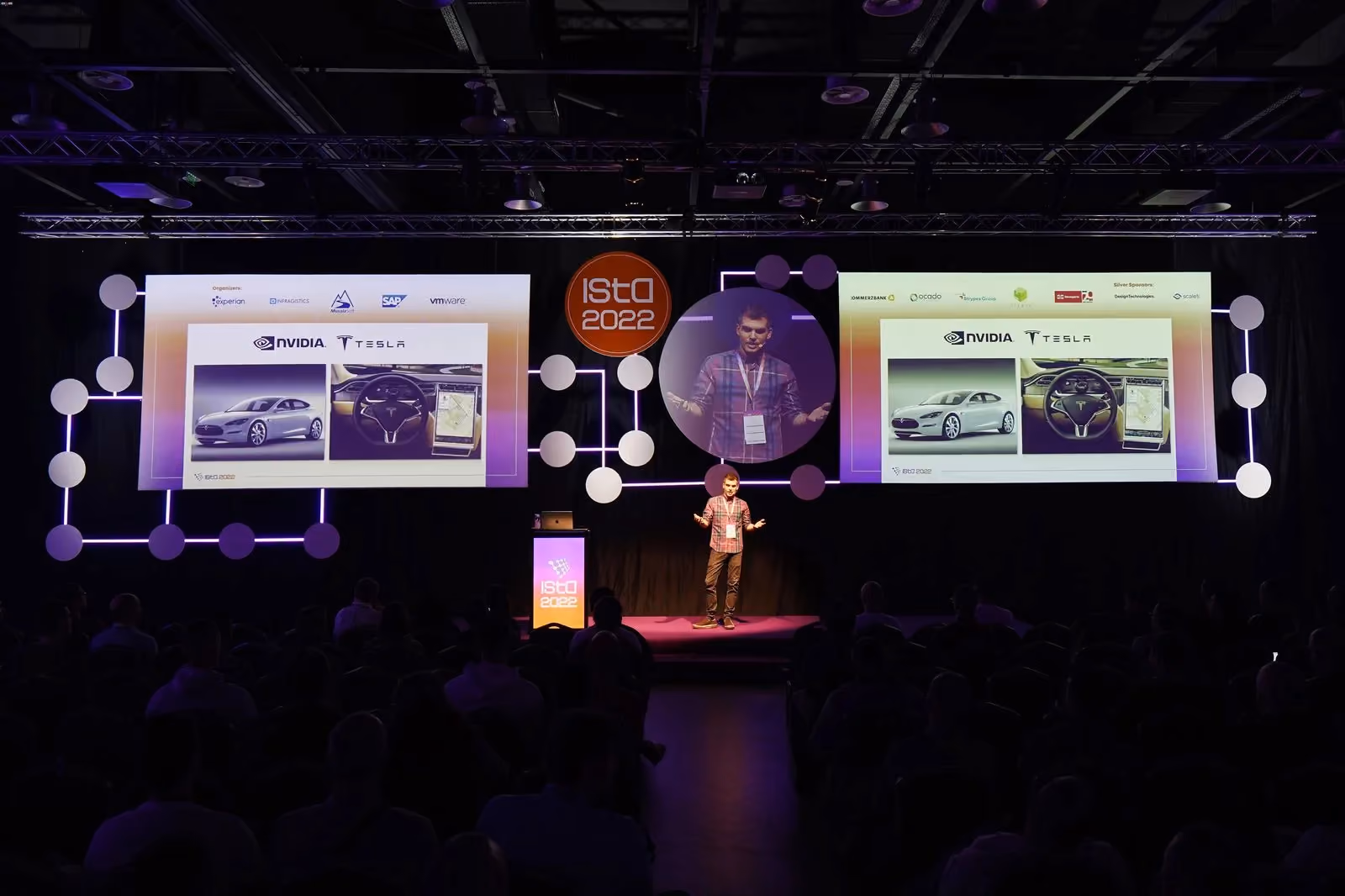 An ISTA 2022 speaker presenting a slide that compares an NVIDIA car and a Tesla interior on twin screens, his live feed in a circular screen above the stage and the seated audience filling the foreground.