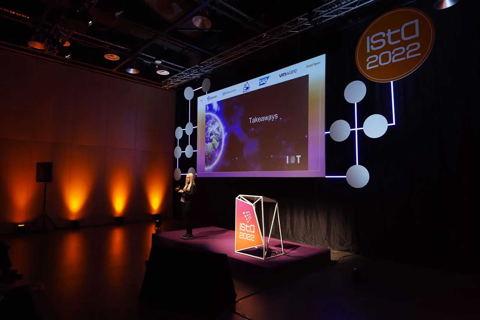 A speaker in black at the edge of the ISTA 2022 stage beside an orange "ISTA 2022" lectern, in front of a screen showing a "Takeaways" slide about IoT.